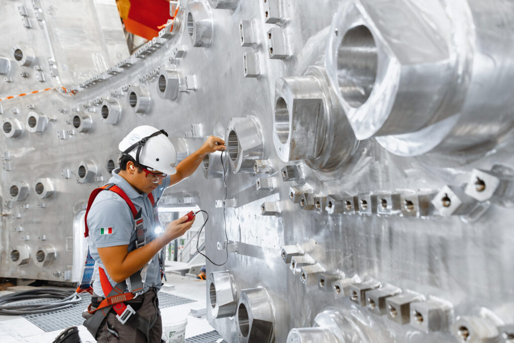 A construction worker in a hard hat regarding a measuring device in his hand with a background of stainless steel wall with openings.