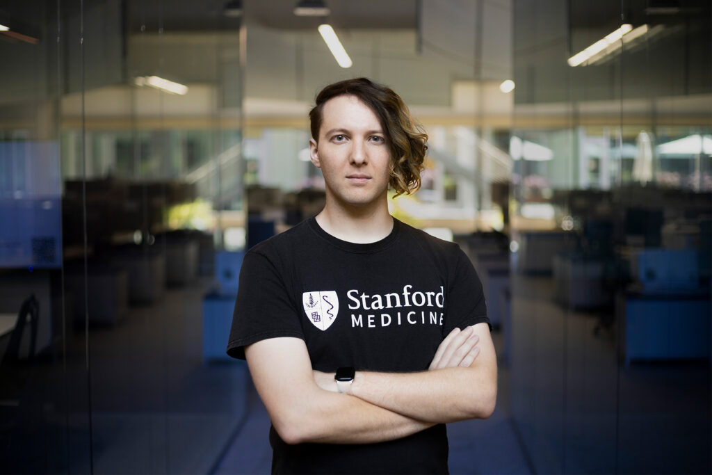A man with an asymmetrical haircut, wearing a black T-shirt that says Stanford Medicine, stands with arms crossed in a glass office space.